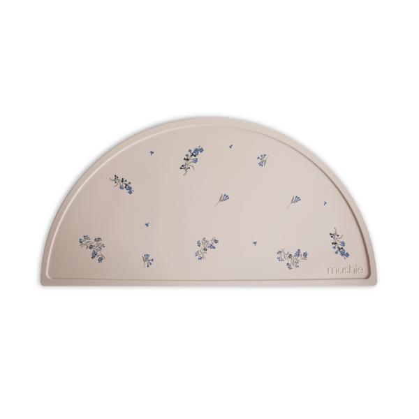 White background with Silicone Place Mat in Lilac by Mushie. Placemat is beige with purple lilacs, made of silicone, and in a semicircle.