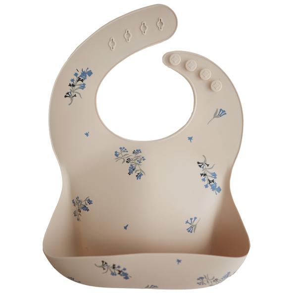 White background with a Silicone Bib in Lilac by Mushie. BIb is beige with purple lilacs, with a deep pocket on the front, and a rounded neck fastener at the back.