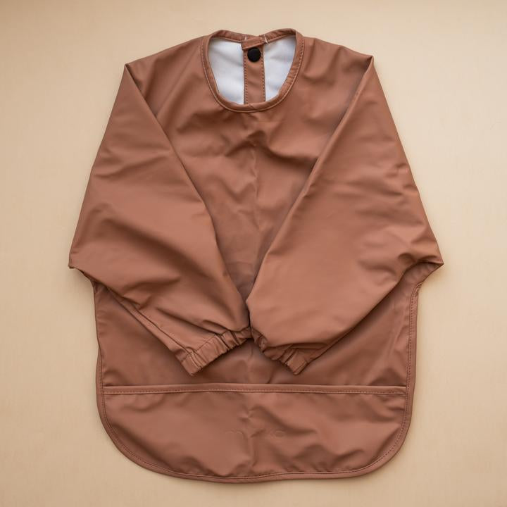Beige background with a Long Sleeve Bib in Cacao by Minika. Bib is dark brown with long sleeves, snaps on the back, and a pouch along the bottom.