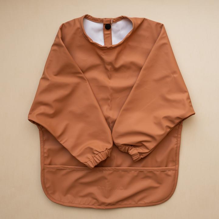 Beige background with a Long Sleeve Bib in Ginger by Minika. Bib is orangey/rust with long sleeves, snaps on the back, and a pouch along the bottom.