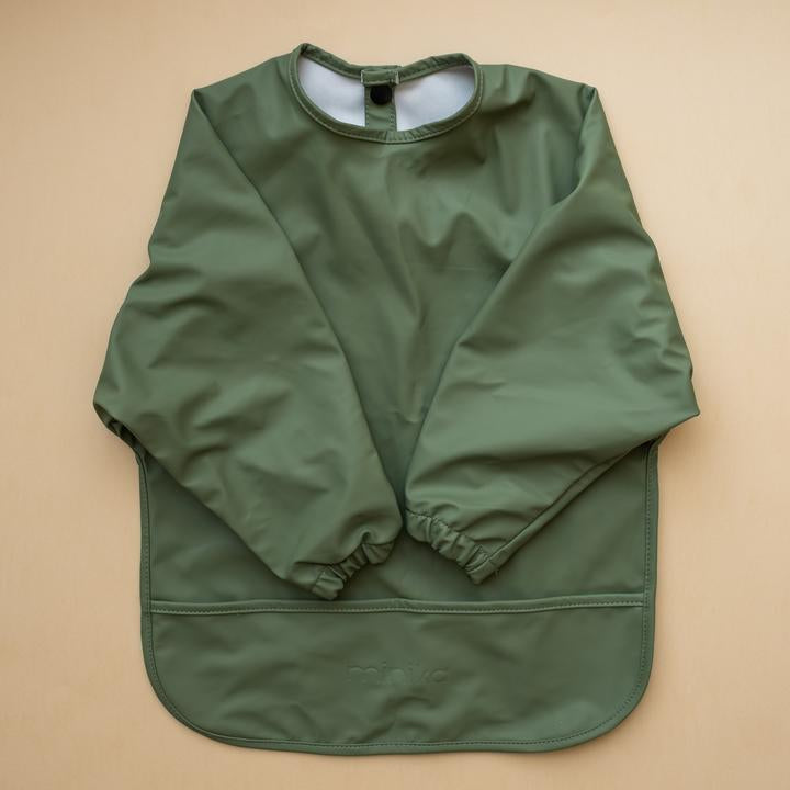 Beige background with a Long Sleeve Bib in Leaf by Minika. Bib is emerald green with long sleeves, snaps on the back, and a pouch along the bottom.