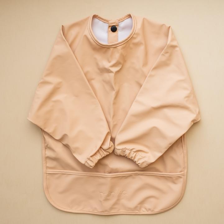 Beige background with a Long Sleeve Bib in Natural by Minika. Bib is ebeige with long sleeves, snaps on the back, and a pouch along the bottom.
