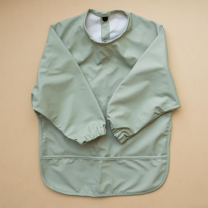 Beige background with a Long Sleeve Bib in Sage by Minika. Bib is sage with long sleeves, snaps on the back, and a pouch along the bottom.