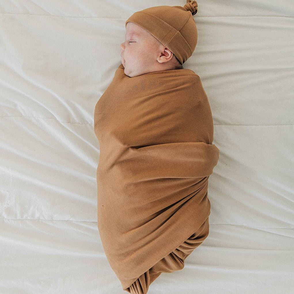 White background with overhead view of baby wrapped in a Mustard Stretch Swaddle by Mebie Baby. Swaddle is mustard, with lots of stretch.