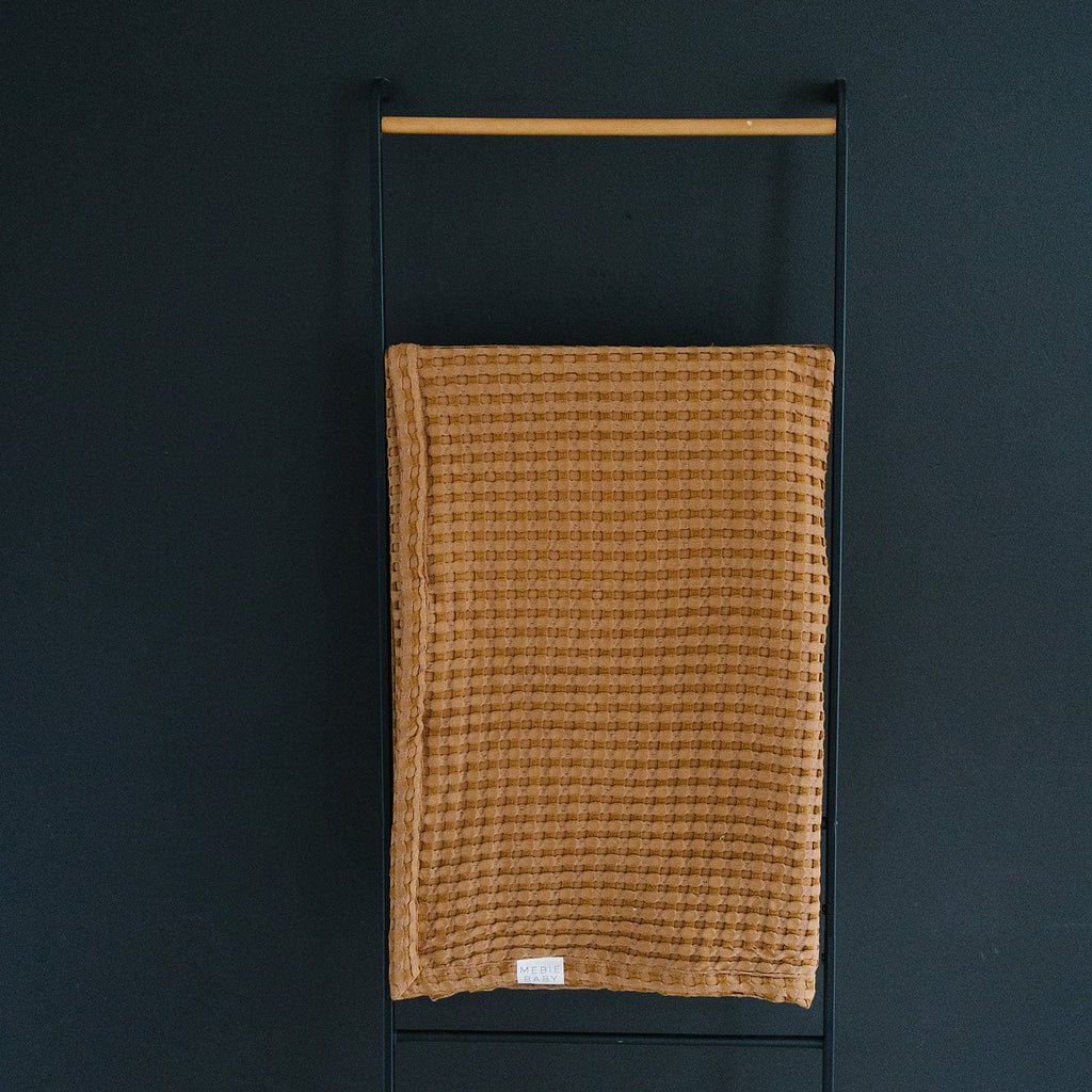 Dark background with a black metal blanket ladder and a Mustard Waffle Quilt by Mebie Baby. Quilt is waffle in a mustard colour.