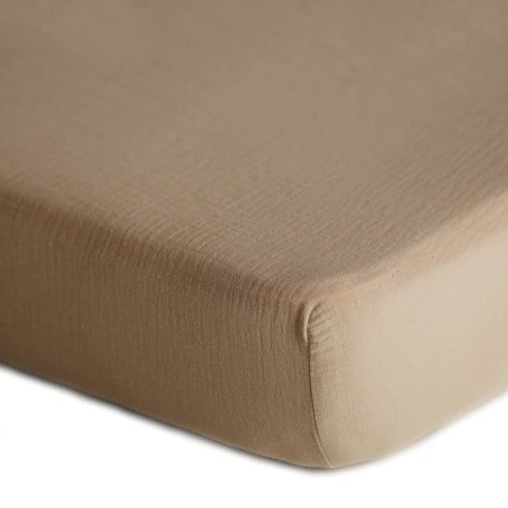 White background with a close up of the corner of a mattress with the Extra Soft Muslin Crib Sheet in Natural by Mushie on it. Natural is a medium grey brown.