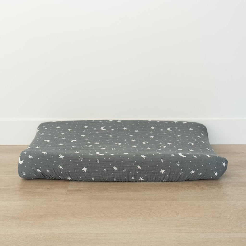 White background, a light wood floor, and a changing pad with a Night Sky Changing Pad Cover by Mebie Baby on it. Changing pad cover is a medium cool blue, with white stars and moons all over.