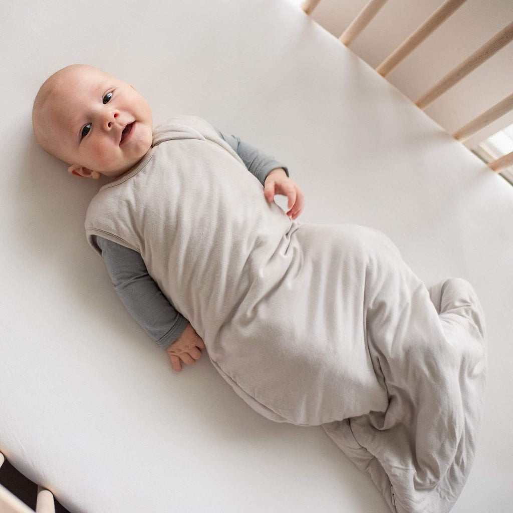 Overhead of baby laying in crib, wearing a Sleep bag 1.0 Tog in Oat by Kyte Baby. Sleep bag is a neutral/grey colour, with a side zipper going all the way to the bottom.