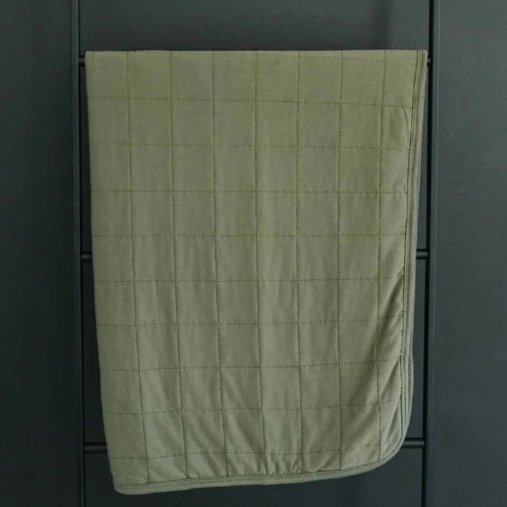 Dark wall with a black metal blanket ladder, and the Olive Bamboo Quilt by Mebie Baby hanging from it. Quilt is an olive colour with quilted details.