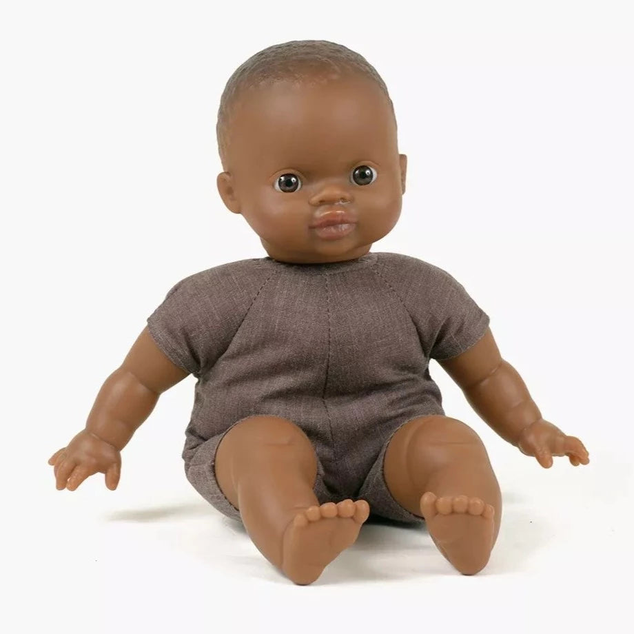 White background with Oscar 28cm Soft Body Doll by Minikane. Doll has black skin, dark eyes, and a dark soft body.