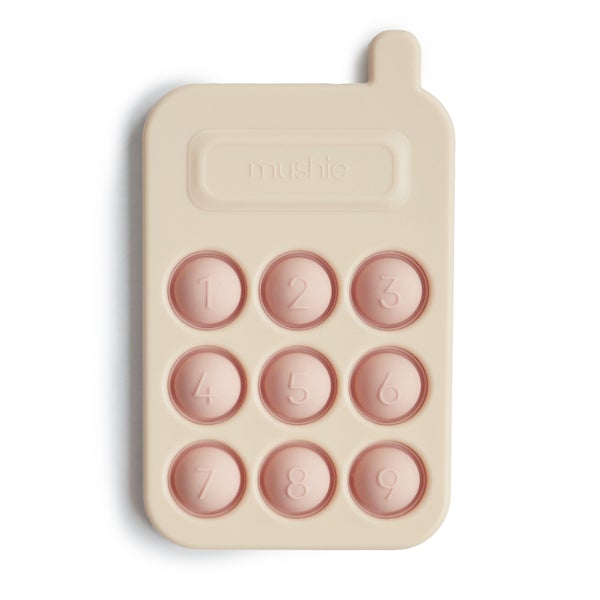 White background with Phone Press Toy in Blush by Mushie. Toy is silicone and looks like a phone, it's cream with little blush coloured number "pop its".
