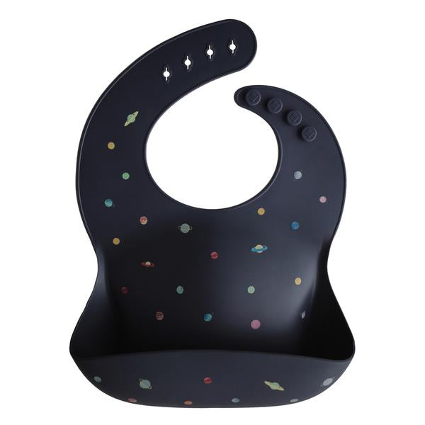 White background with a Silicone Bib in Planets by Mushie. Bib is a dark blue with multicoloured planets, with a deep pocket on the front, and a rounded neck fastener at the back.