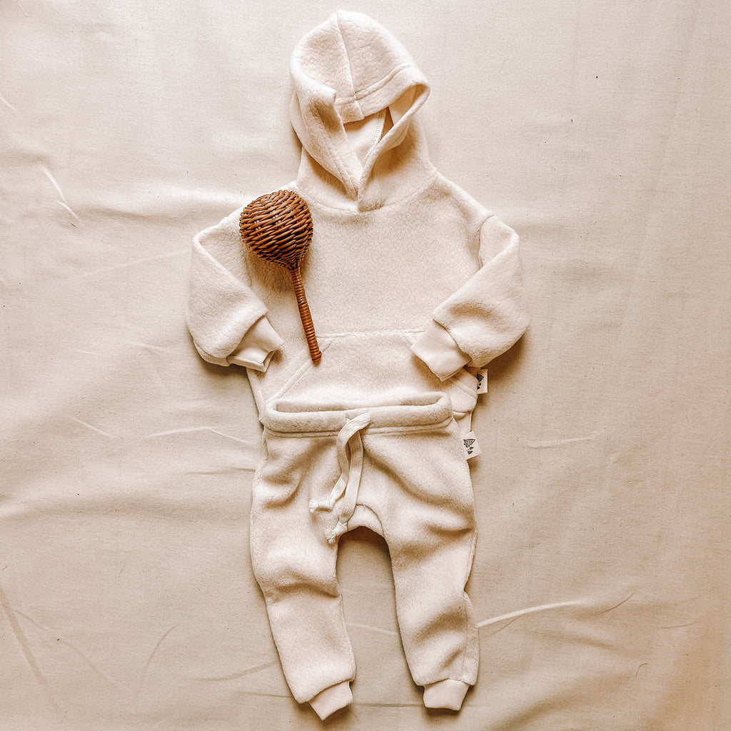 Polar Bear hoodie and pant by Petit Nordique, with a rattan wood baby rattle. Laid on a flat beige surface. 