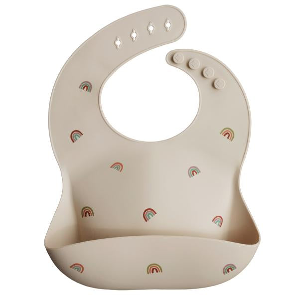White background with a Silicone Bib in Rainbows by Mushie. Bib is a cream colour with multicolour rainbows, with a deep pocket on the front, and a rounded neck fastener at the back.