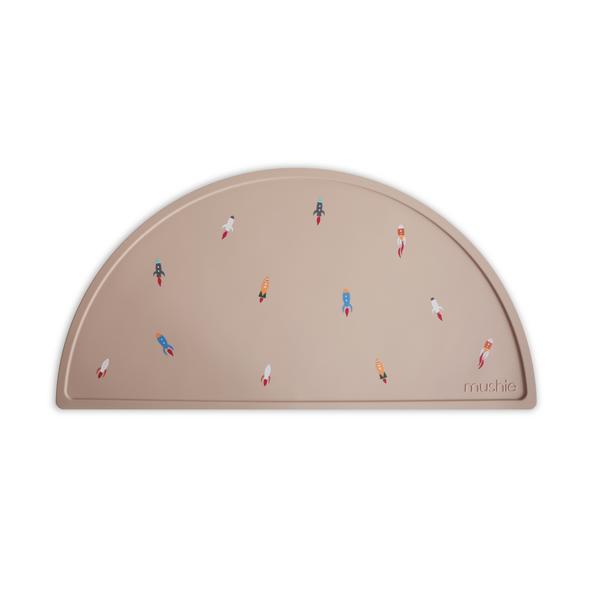 White background with Silicone Place Mat in Rocket Ship by Mushie. Placemat is tan with colourful rocket ships, made of silicone, and in a semicircle.