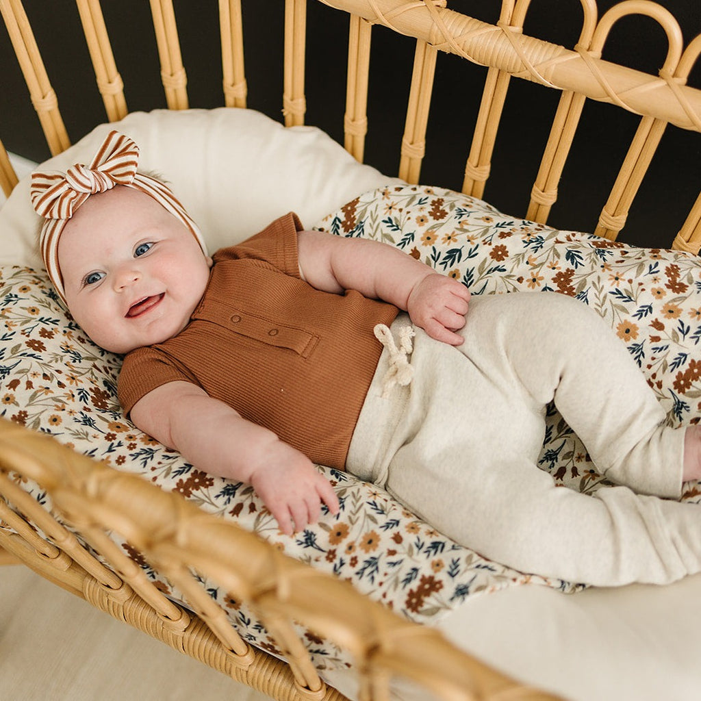 Rust Organic Bodysuit by Mebie Baby with a baby wearing oatmeal coloured pants with a Harvest Floral Swaddle by Mebie Baby in a wood bassinet. 