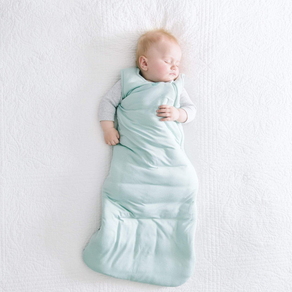 White background with baby sleeping, laying on back, wearing a Sleep Bag 1.0 Tog in Sage by Kyte Baby. Sleep bag is a light green colour, with a side zipper going all the way down to the bottom.