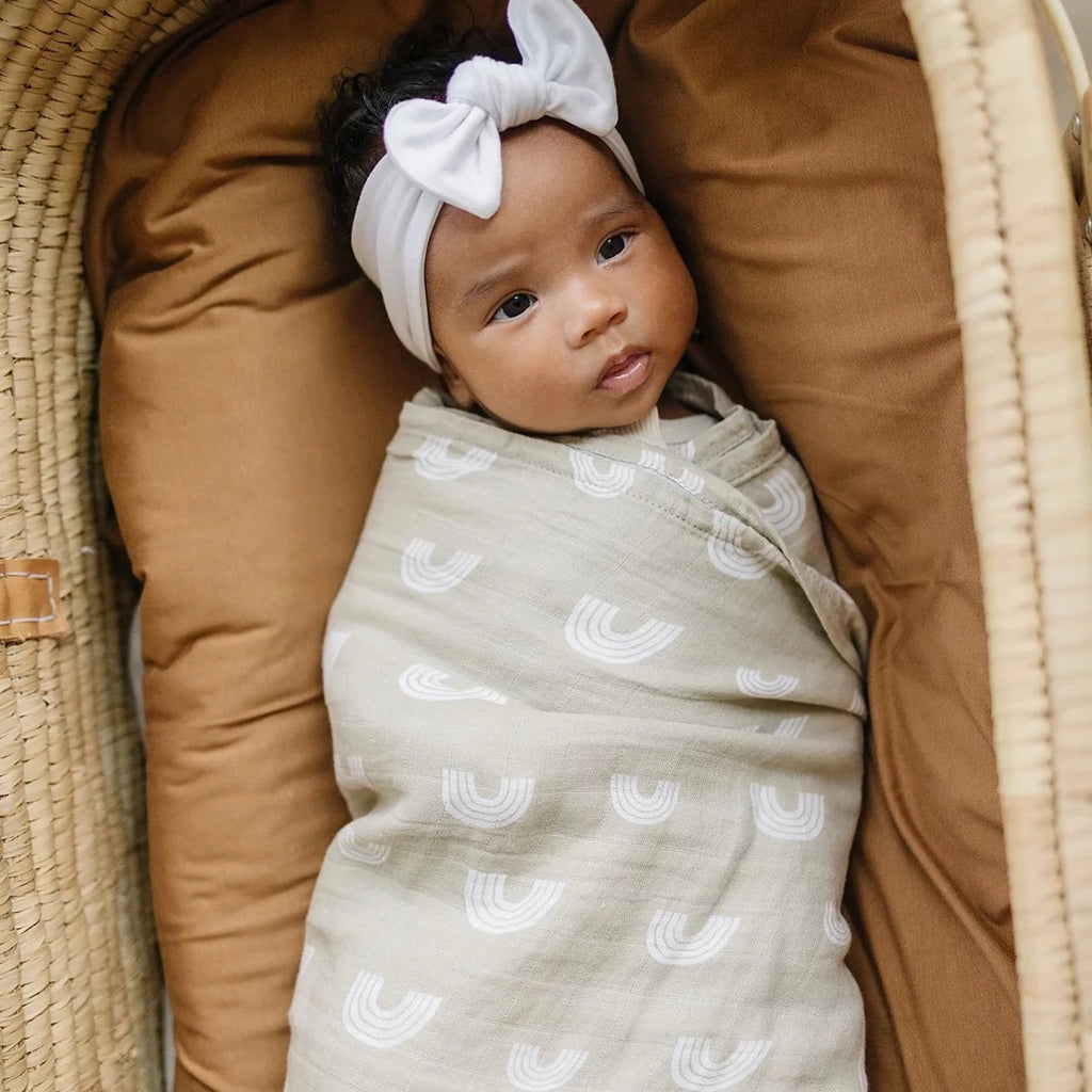 Overhead view of a baby girl laying in a rattan bassinet, wrapped up in the Sand Rainbow Muslin Swaddle by mebie Baby.