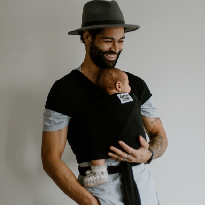 Model wearing the Sawyer Wrap Carrier by Beluga Baby, while holding a baby in front of a grey wall. Model wearing a fedora. 