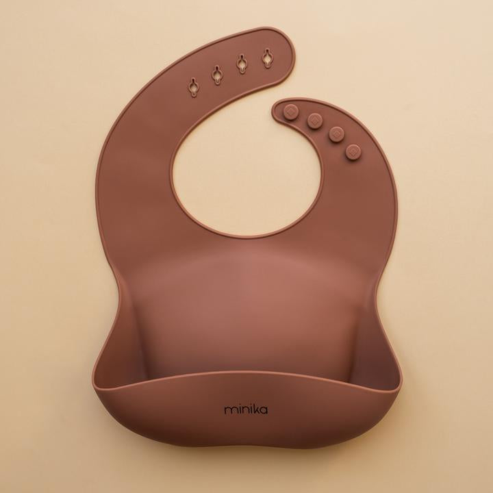 Beige background with a Silicone Bib in Cacao by Minika. Bib is dark brown colour, and has a pouch on the front.