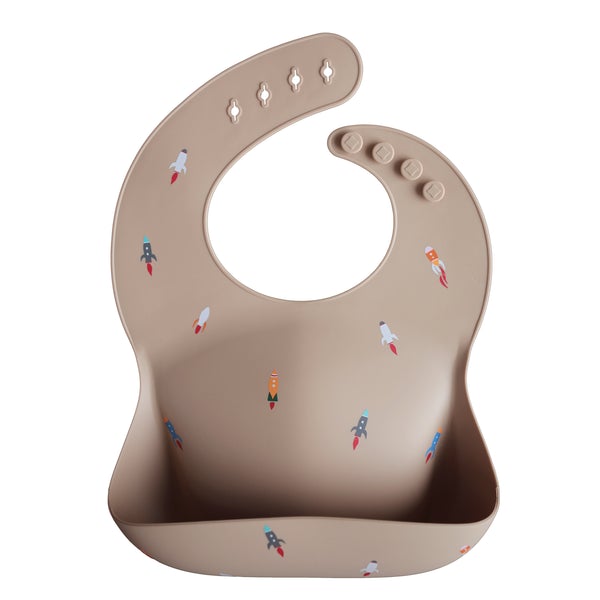 White background with a Silicone Bib in Rocketship by Mushie. Bib is tan with colourful rocket ships, with a deep pocket on the front, and a rounded neck fastener at the back.