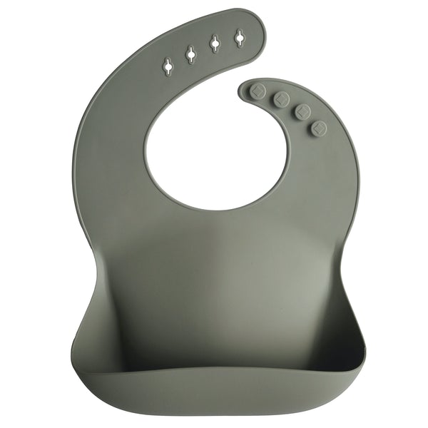 White background with a Silicone Bib in Silver Sage by Mushie. Bib is a deep grey olive colour, with a deep pocket on the front, and a rounded neck fastener at the back.