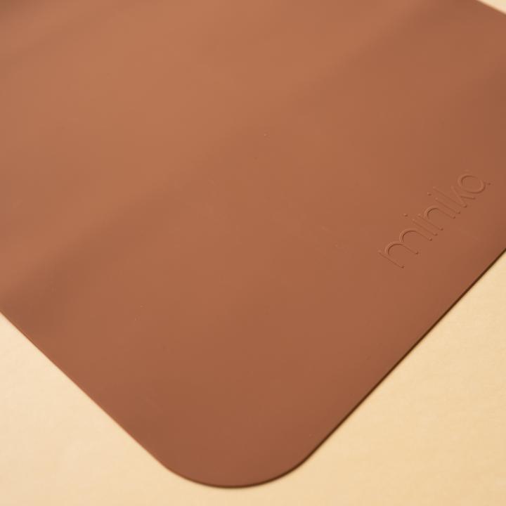 Beige background with a Silicone Placemat in Cacao by Minika. Placemat is square silicone, in a dark brown colour.