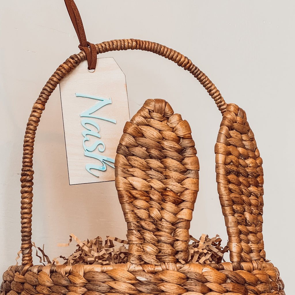 Customized name tag for Nash in Sky by Petit Nordique Boutique hanging on an Easter Bunny Basket in front of a beige wall. 
