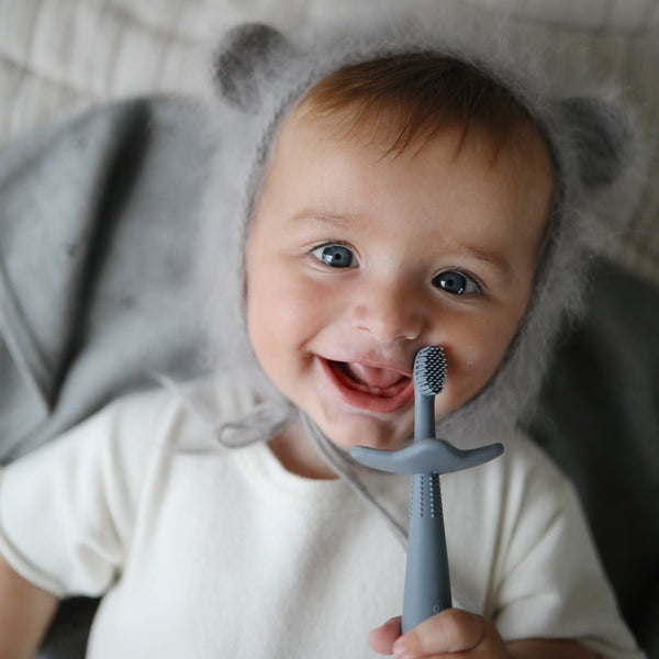 Overhead view of a baby laying down, smiling, with a grey background, and he's holding a Star Training Toothbrush by Mushie.
