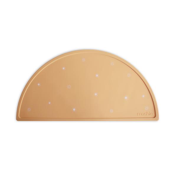 White background with Silicone Place Mat in Sun Orange by Mushie. Placemat is orangey yellow with white suns, made of silicone, and in a semicircle.