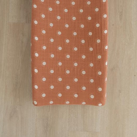 Overhead view of a light wood floor and a change pad with the Sunshine Changing Pad Cover by Mebie Baby on it. Cover is orange with white suns.