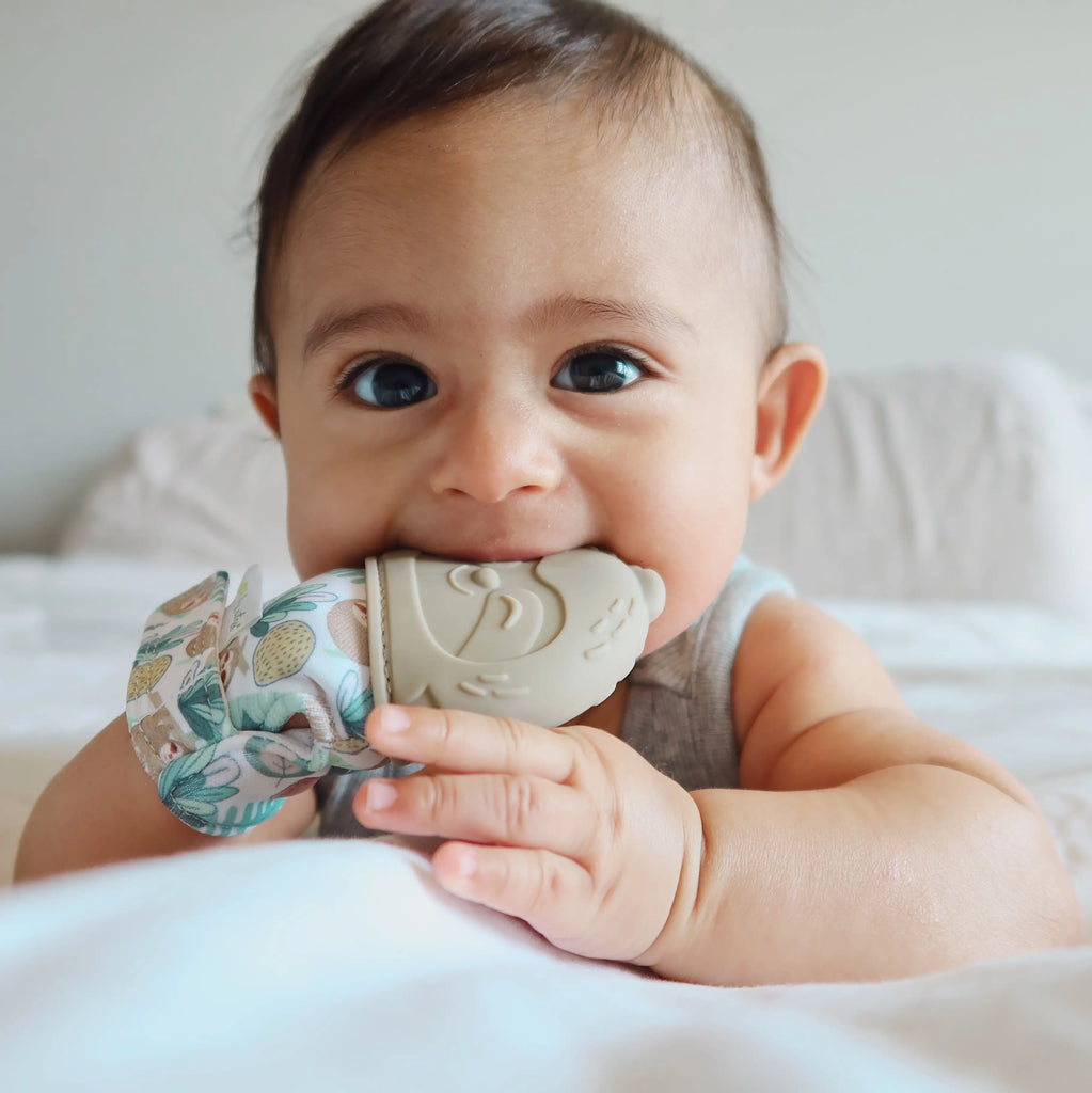 White background, and baby laying on their tummy on a bed with the Itzy Mitt™ Silicone Teething Mitt in Sloth by Itzy Ritzy on their hand. Mitt has a beige silicone sloth, and the glove part is a crinkly fabric in white with a jungle sloth print.
