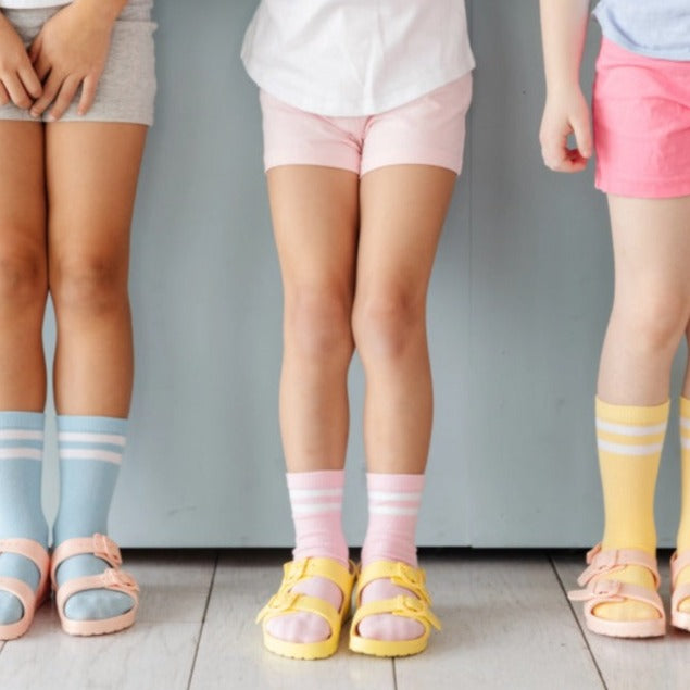 Close up of 3 girls standing against a blue wall, wearing Pastel Striped Midi 3-Pair Pack by Little Stocking Co. Colours are blue, pink, and yellow.