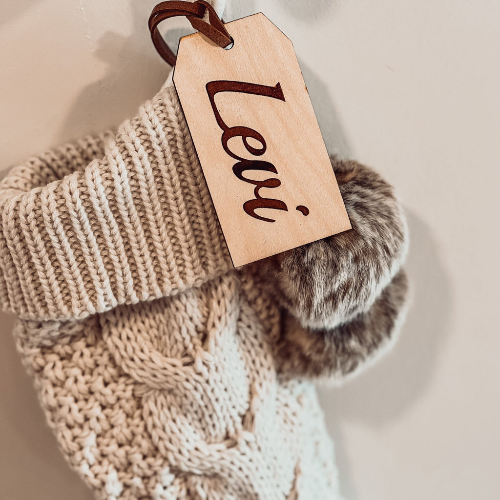 Customized Wooden Tag engraved with the name Levi by Petit Nordique, hanging on a beige Christmas stocking with faux fur pompoms in front of a beige wall. 