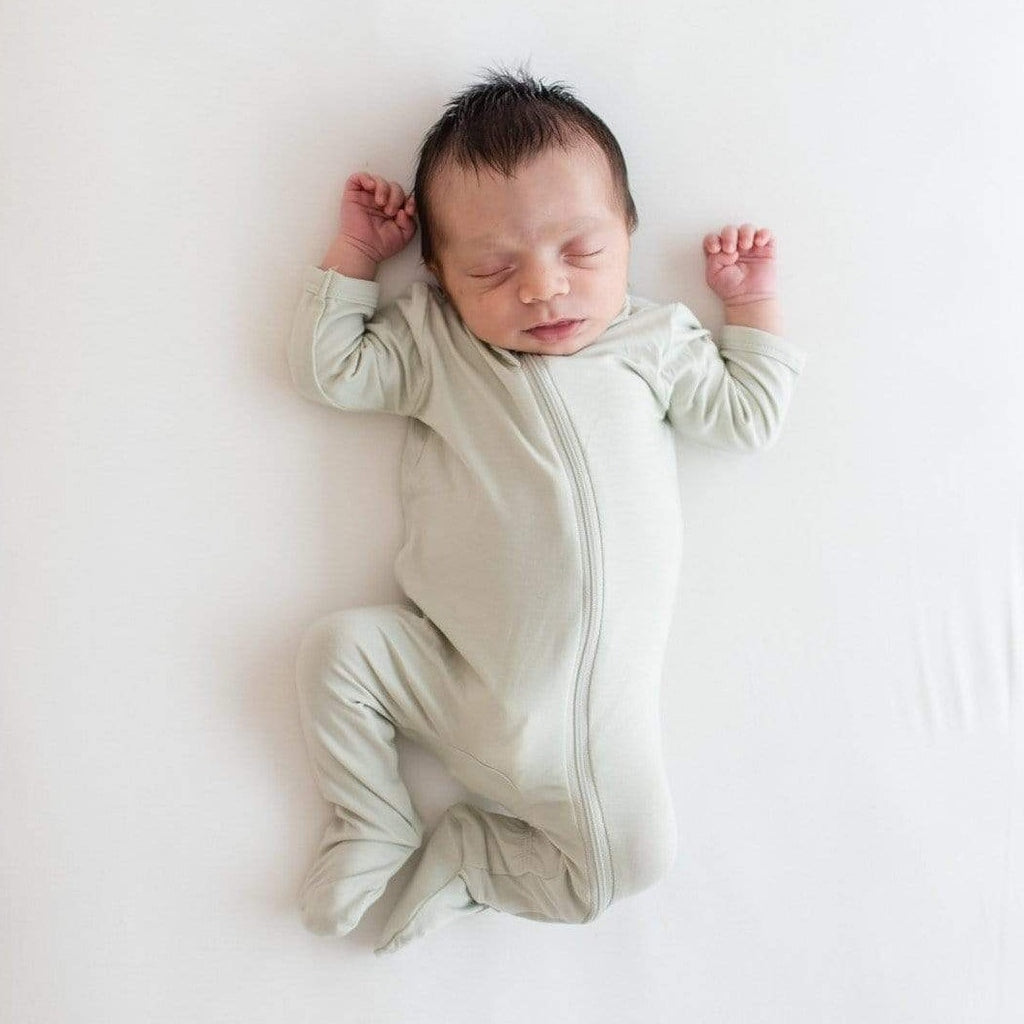 White background, overhead view of baby sleeping, wearing a Zippered Footie in Aloe by Kyte Baby. Zippered Footie is a very pale green with a zipper that runs all the way down the front.