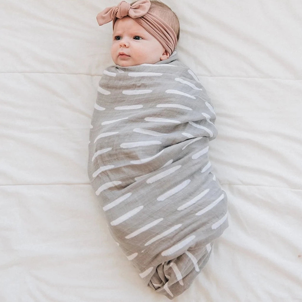 Overhead view of baby laying down, wrapped up in a Grey Dash Muslin Swaddle by Mebie Baby. Swaddle is a medium grey with white dashes all over.