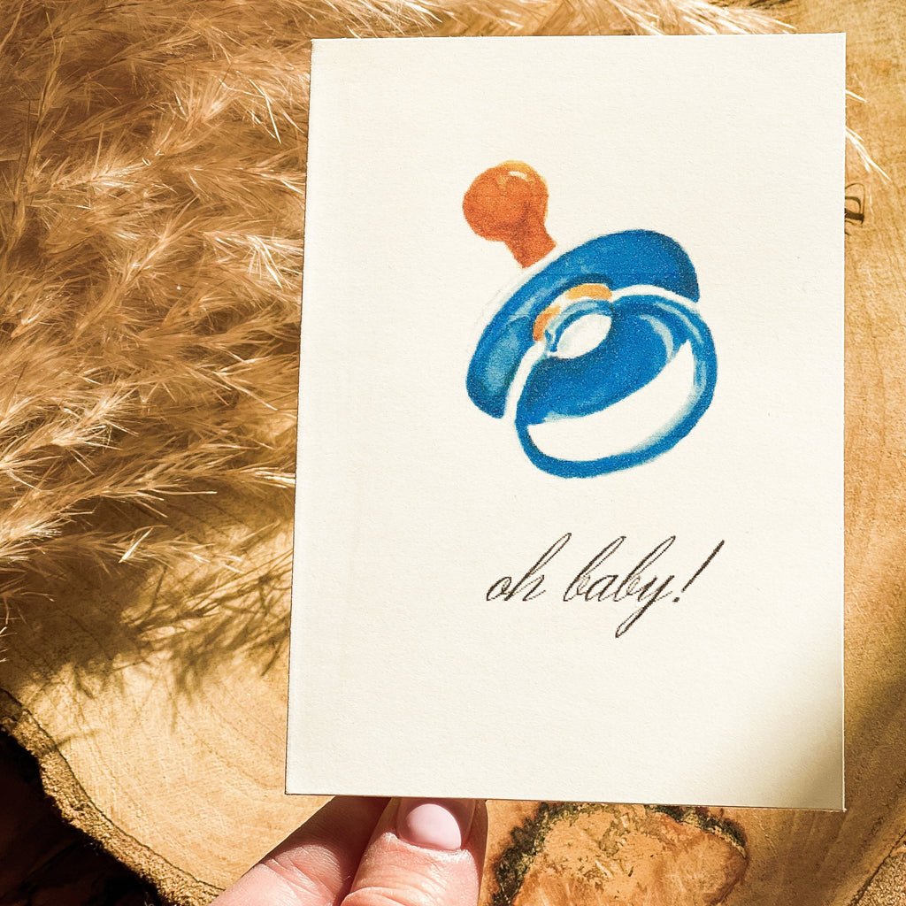 Oh Baby! | Blue Paci card by Parkes & Bash, being held in front of wood and grass pampas with a hand holding it up. 