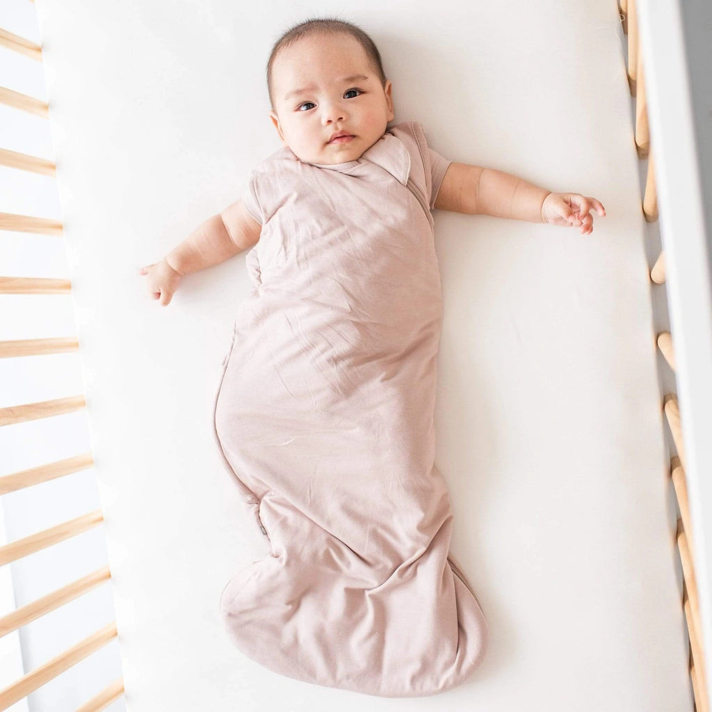 Overhead shot of baby laying on her back in crib, wearing a Sleep Bag .5 Tog in Sunset by Kyte Baby. Sleep Bag is soft, with a side zipper, and is a neutral pink colour.