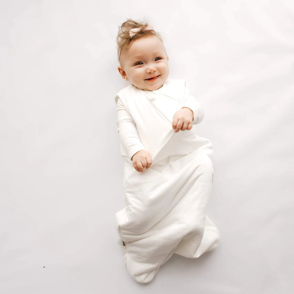 White background with baby laying down, wearing a Sleep Bag 2.5 Tog in Cloud by Kyte Baby. Sleep bag is white, with a zipper all the way down to the bottom.