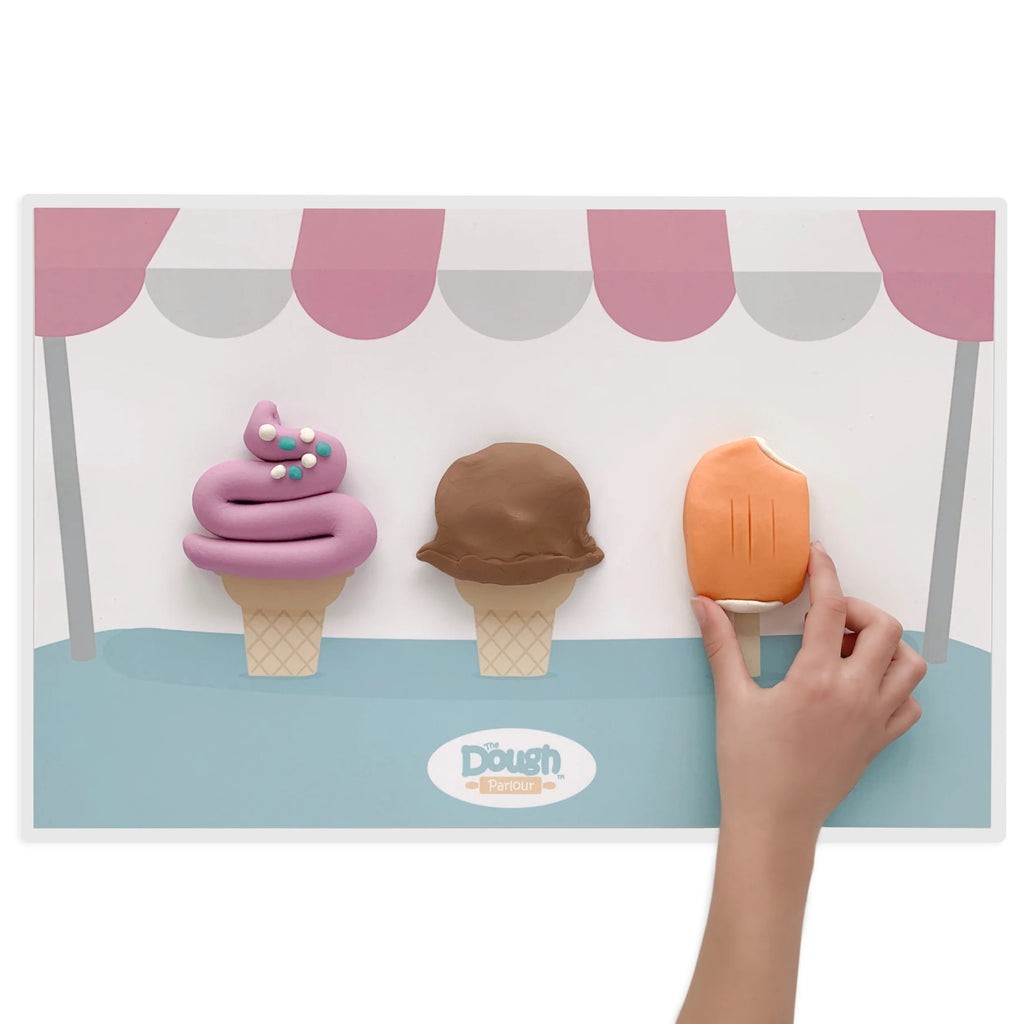 White background with Playmat by Dough Parlour. Playmat is of 2 ice cream cones, and a popsicle stick.. hand has molded dough to look like ice cream, and a popsicle.
