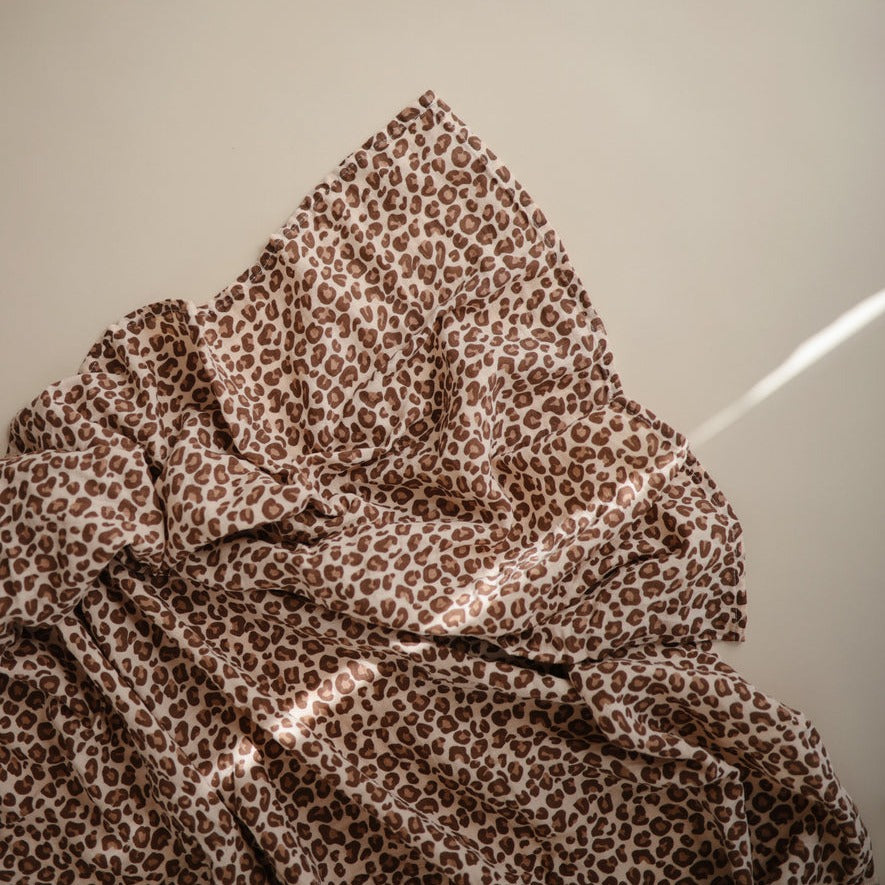 Muslin Swaddle Blanket Organic Cotton in Leopard by Mushie laid on a beige surface. 