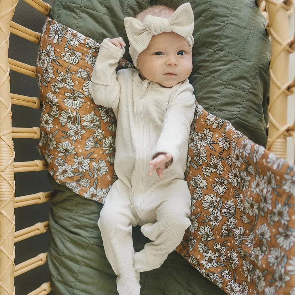 Magnolia Muslin Swaddle by Mebie Baby baby girl laying in wooden wicker bassinet 