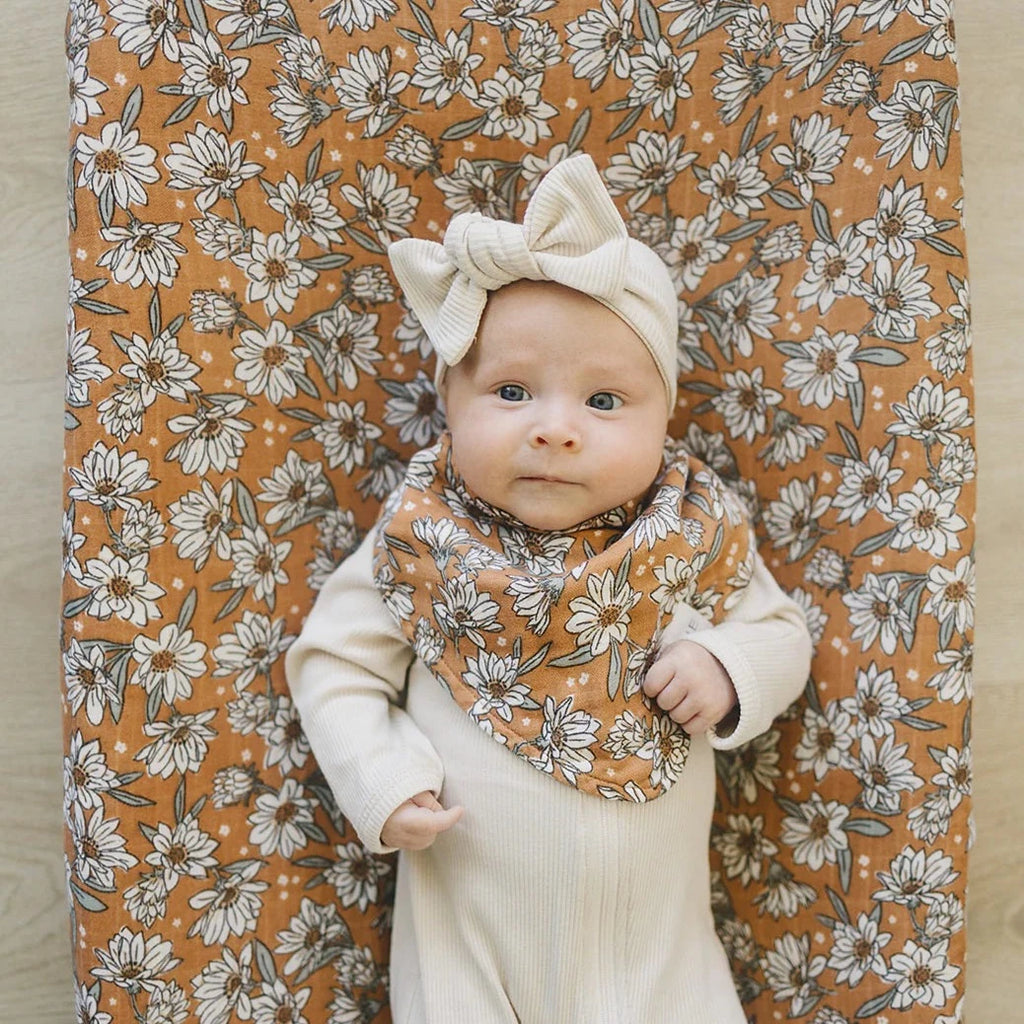 Magnolia Bib by Mebie Baby drool bib on baby girl laying on mattress with oatmeal ribbed headband bow