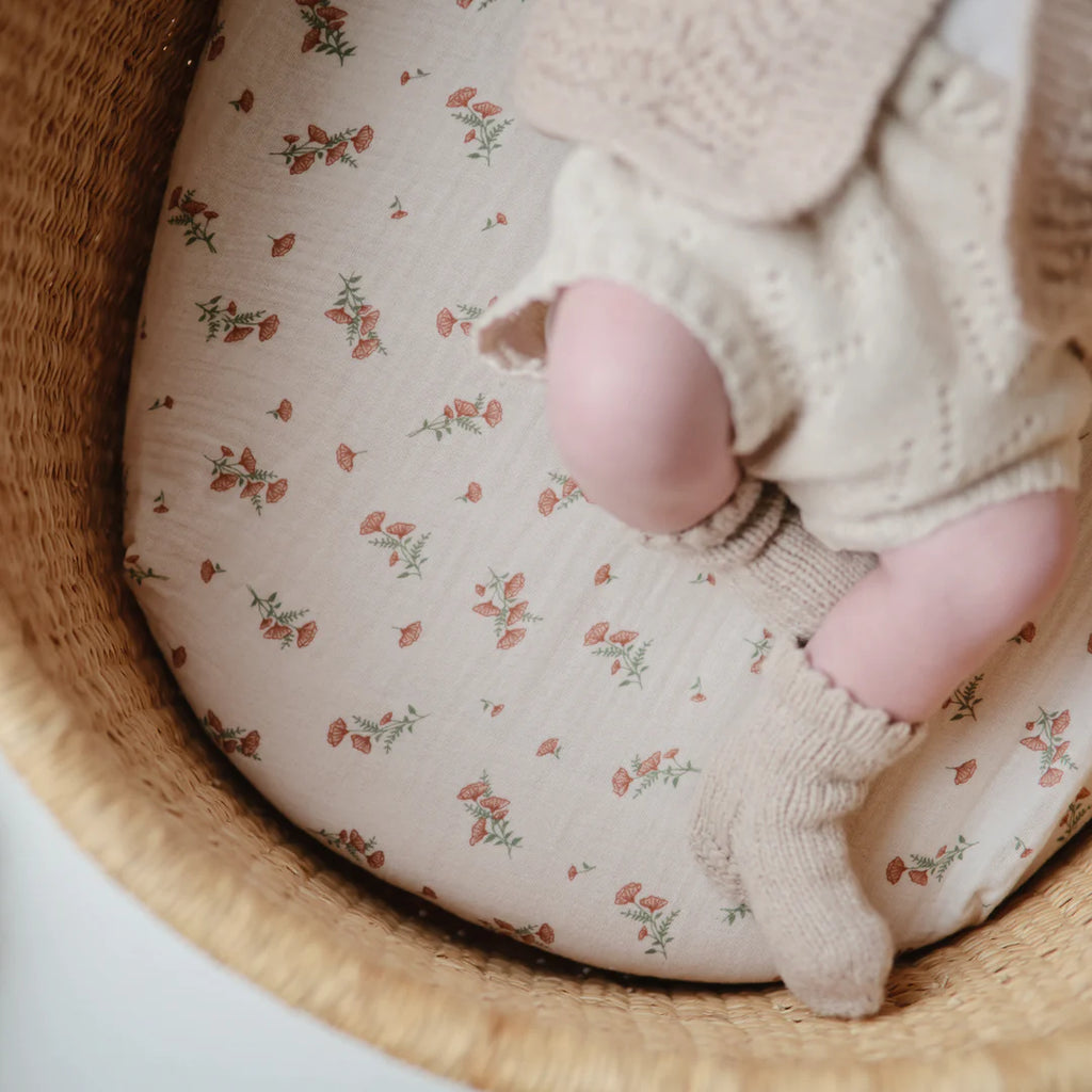 Muslin Bassinet Sheet | Pink Flowers by Mushie baby legs in wicker bassinet wearing knit socks