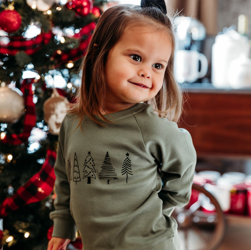 little girl in front of christmas tree with green trees sweatshirt