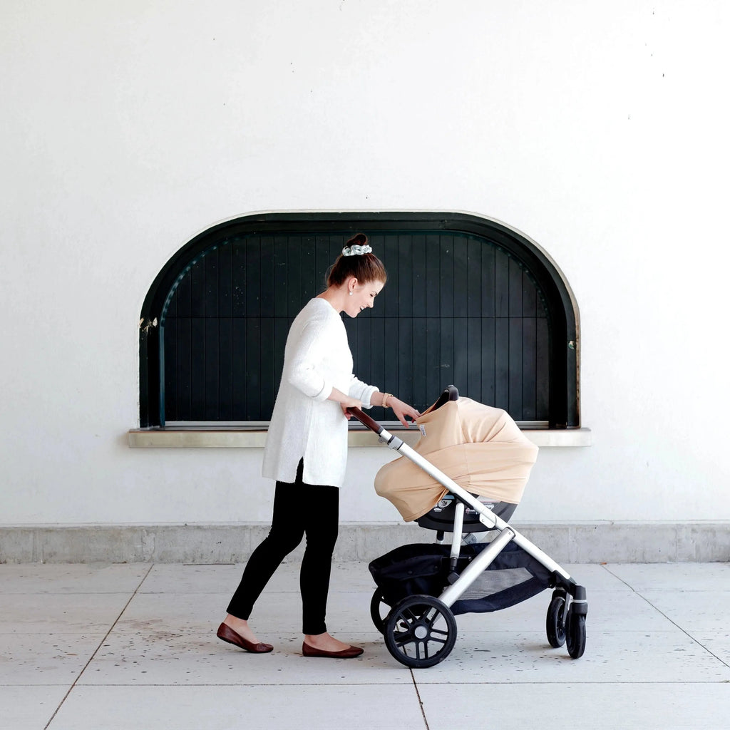 White wall with a black window arch, and a mama pushing her baby in a stroller with The OVer Cover in Iced Coffee by The OVer Company on her carseat. This cover is a yellowy brown colour, with a white tag with black text that says "OV" with black greenery.