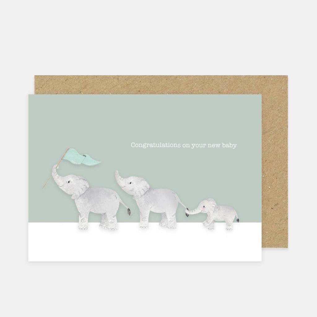Light background with a 'Congratulations on your new baby' Card by Little Roglets. Card is on top of a craft paper envelope, and card is a grey/green with 3 Elephants in a line, the front one has a flag, and the back one is holding the middle ones tail with their trunk.