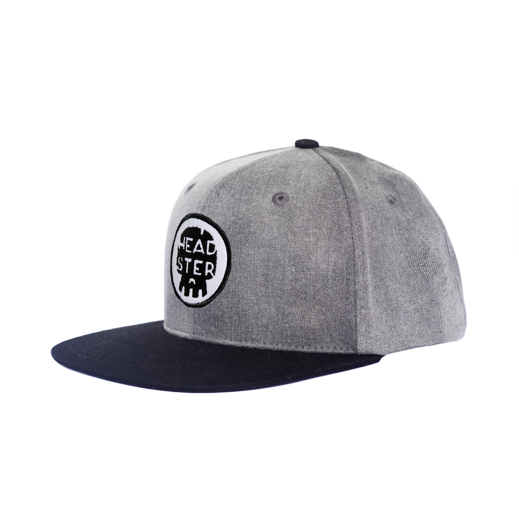 White background with Dark G-NZ Snapback Hat by Headster. Snapback has a black brim, with a dark grey hat, and a patch on the front that says "Headster".