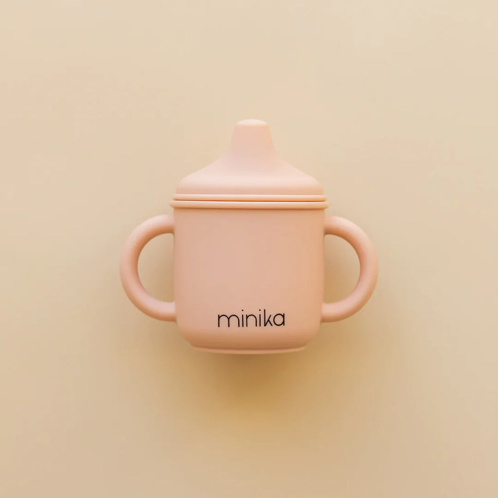 Beige background with a Silicone Sippy Cup in Blush by Minika. Sippy cup is made of silicone, with 2 handles, a spouted lid, and the colour is blush and says “minika” in black.