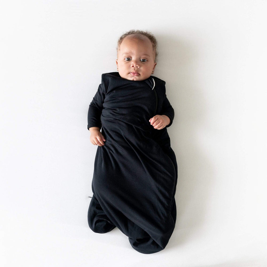 White background with overhead view of baby, laying down, wearing a Sleep Bag 2.5 Tog in Midnight by Kyte Baby. Sleep bag is black, with a side zipper going all the way around the bottom.